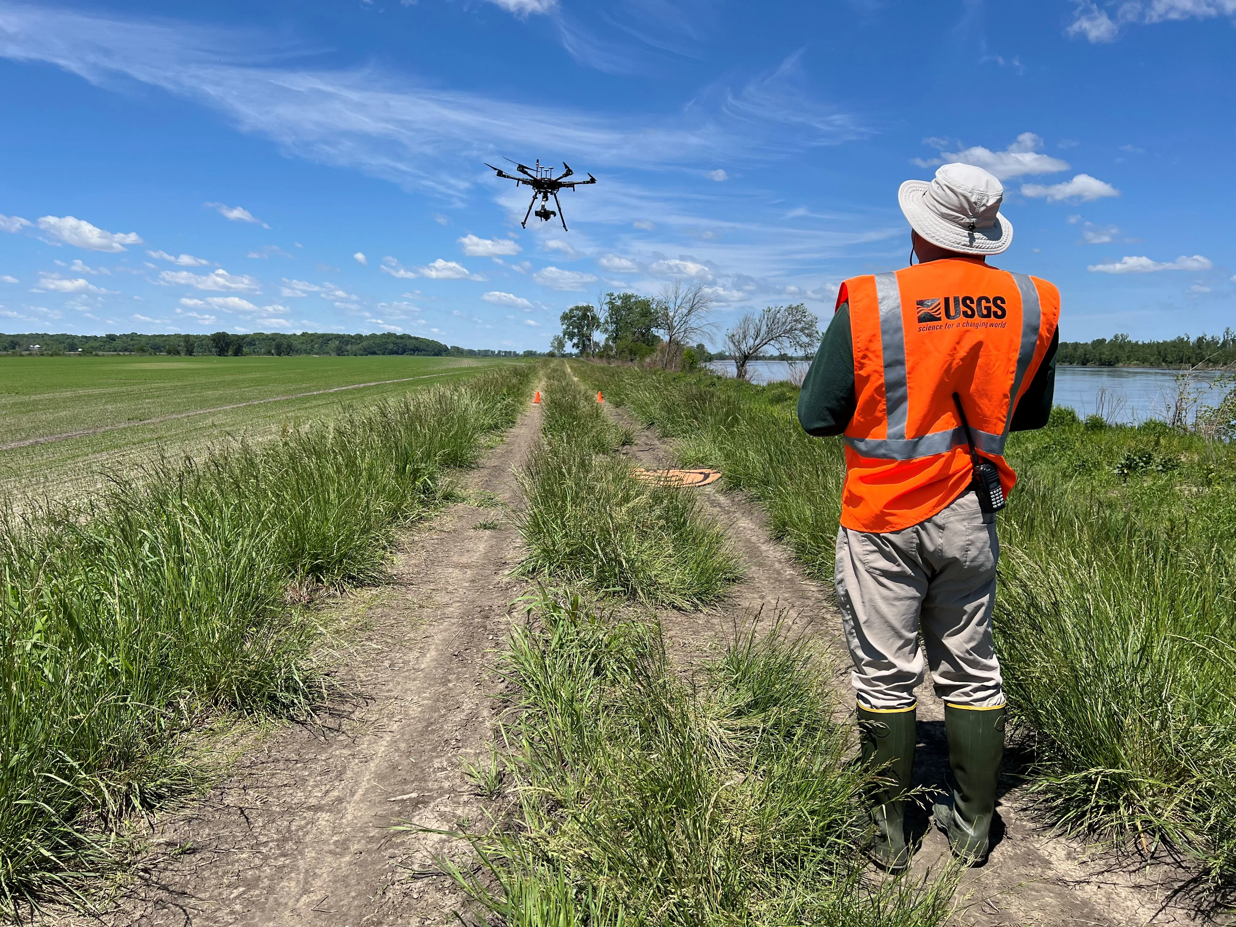 A multirotor aircraft carrying a suspended payload flies above a levee while an operator in an orange USGS vest watches from beside the river.