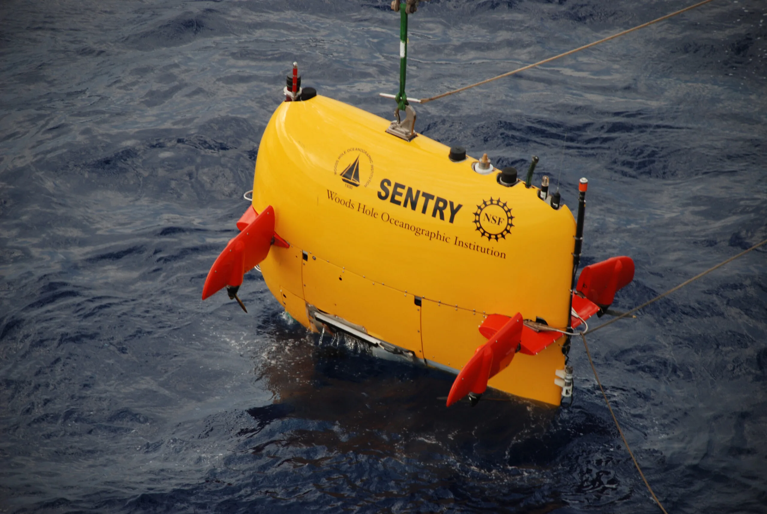 Yellow Sentry autonomous underwater vehicle with red fins suspended at the sea surface by a lifting line