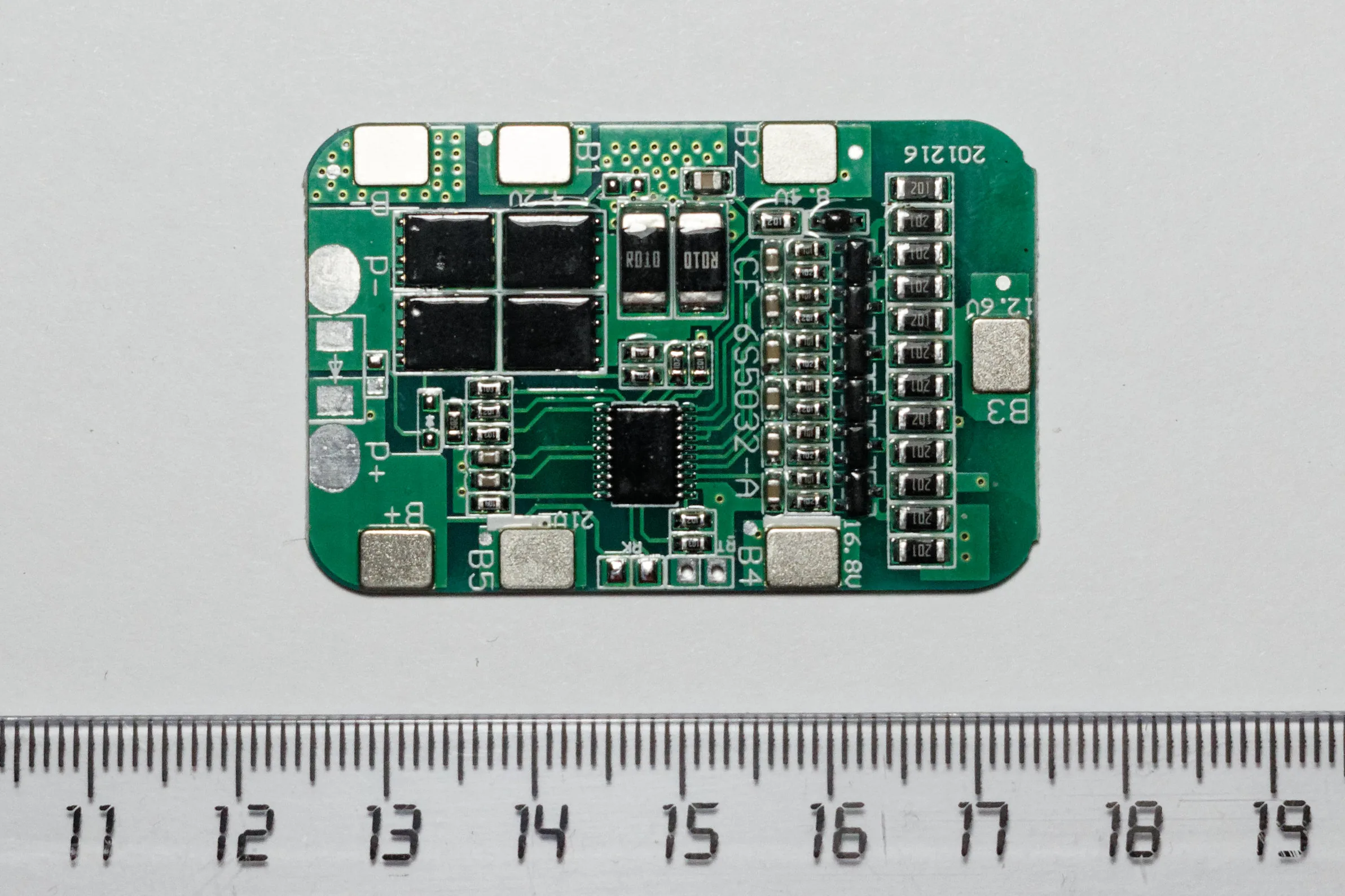 Green battery management circuit board with black components, labeled connection pads, and a ruler below it.