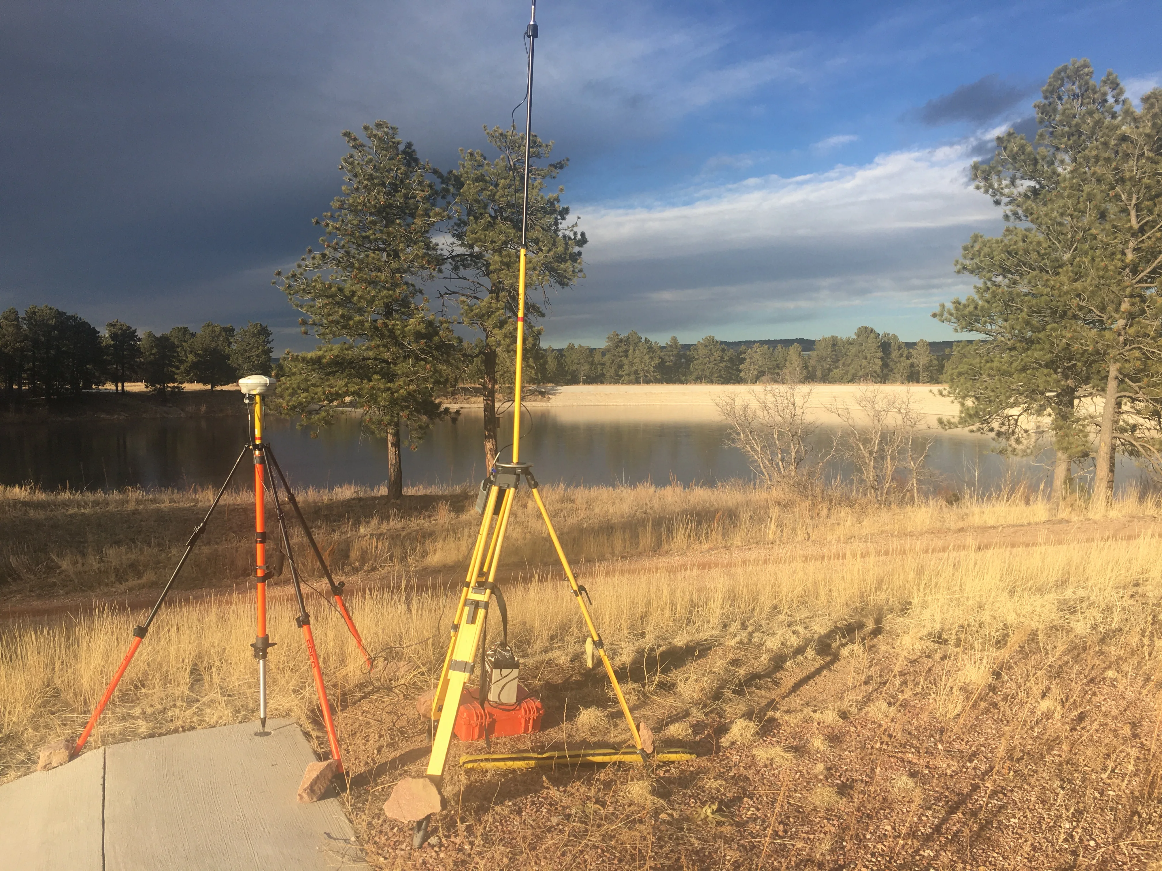 Tripod-mounted GNSS surveying equipment and a tall radio antenna beside a tree-lined reservoir.
