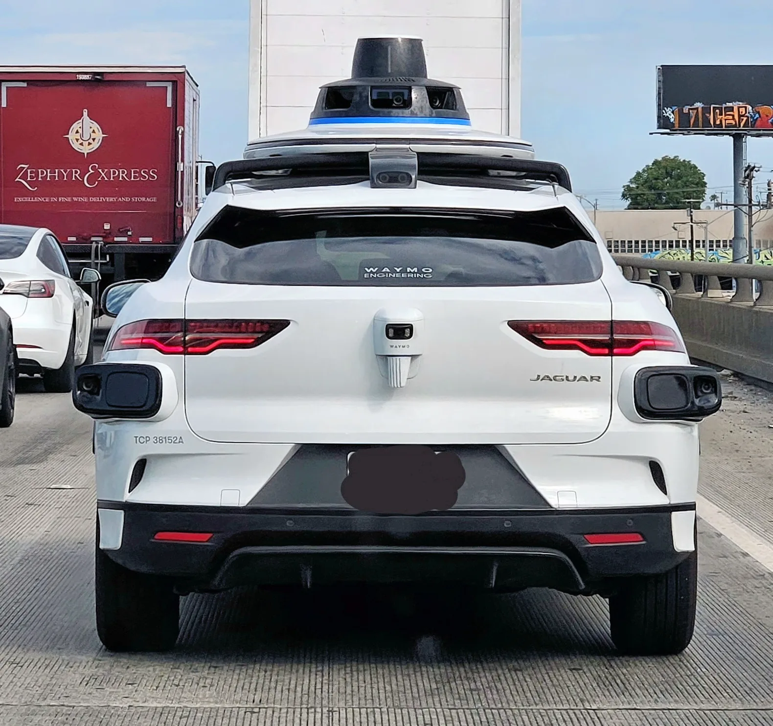 Rear view of a white Waymo Jaguar in road traffic, with roof-mounted sensors and additional rear sensor housings.