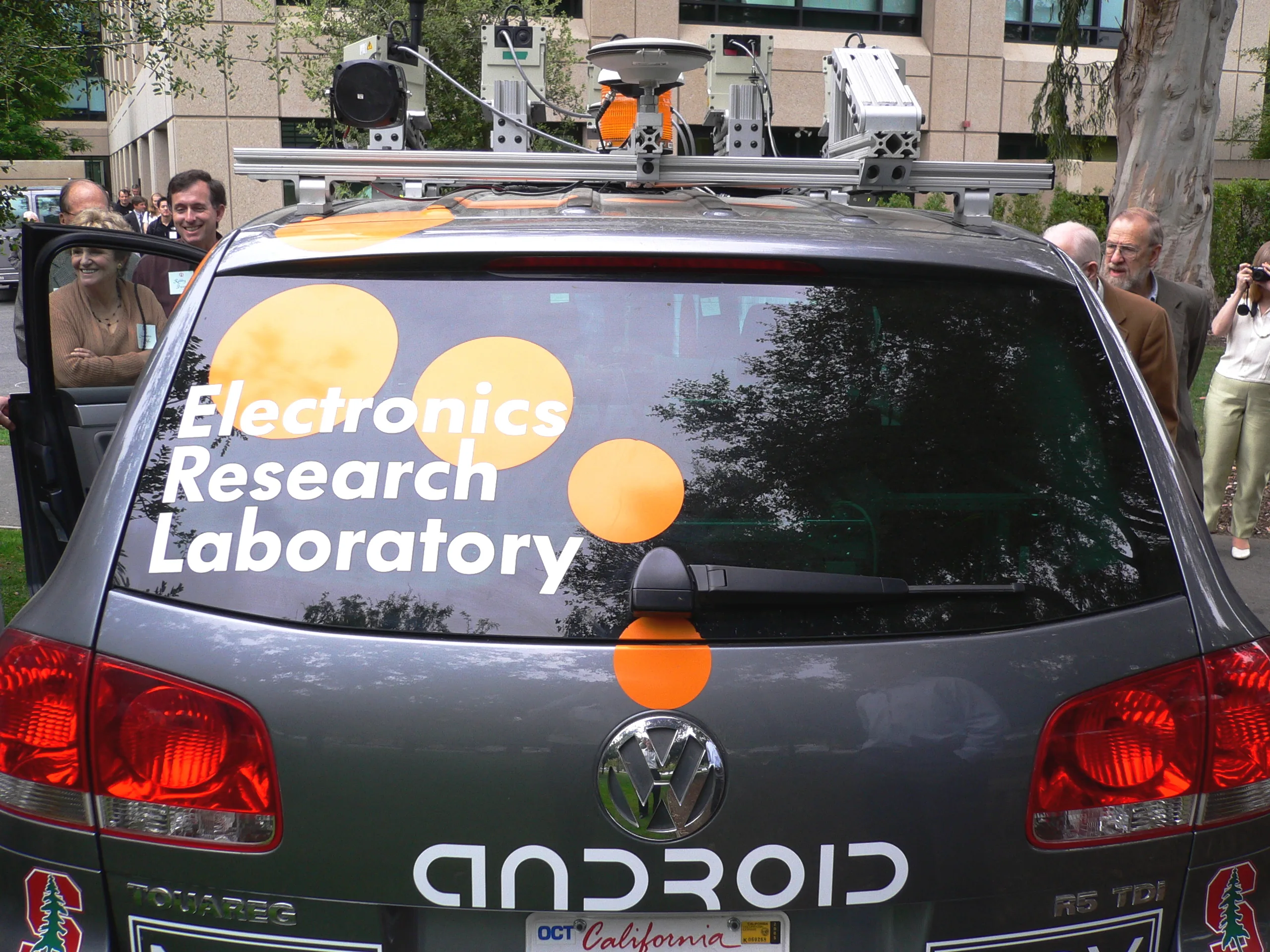Rear view of Stanford autonomous vehicle Stanley with sensors mounted on a roof rack and people standing beside it.
