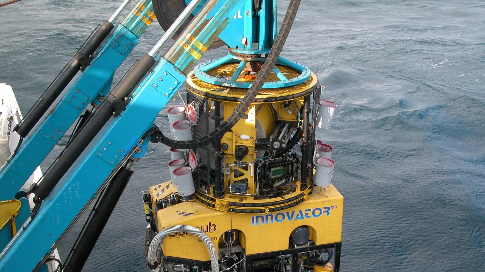 A cylindrical tether management assembly sits above a yellow remotely operated vehicle suspended beside a vessel over the sea.