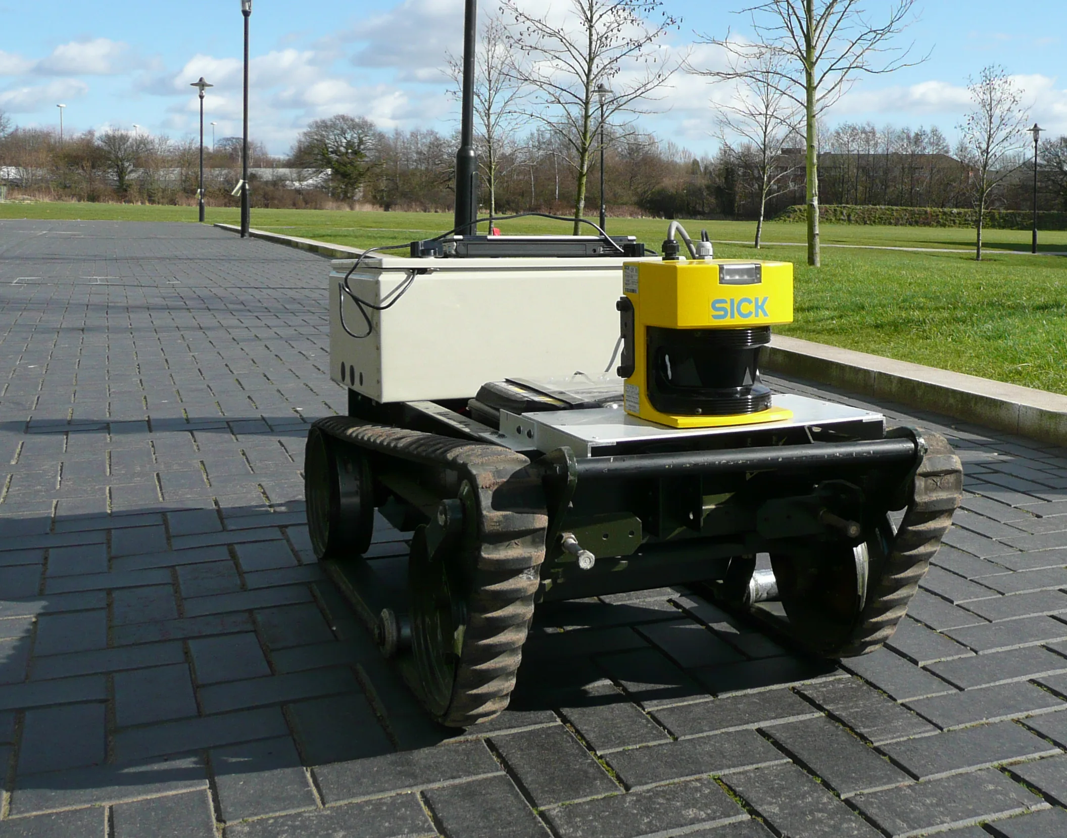 A wheeled mobile robot with a yellow lidar scanner mounted above its front wheels on a paved outdoor path