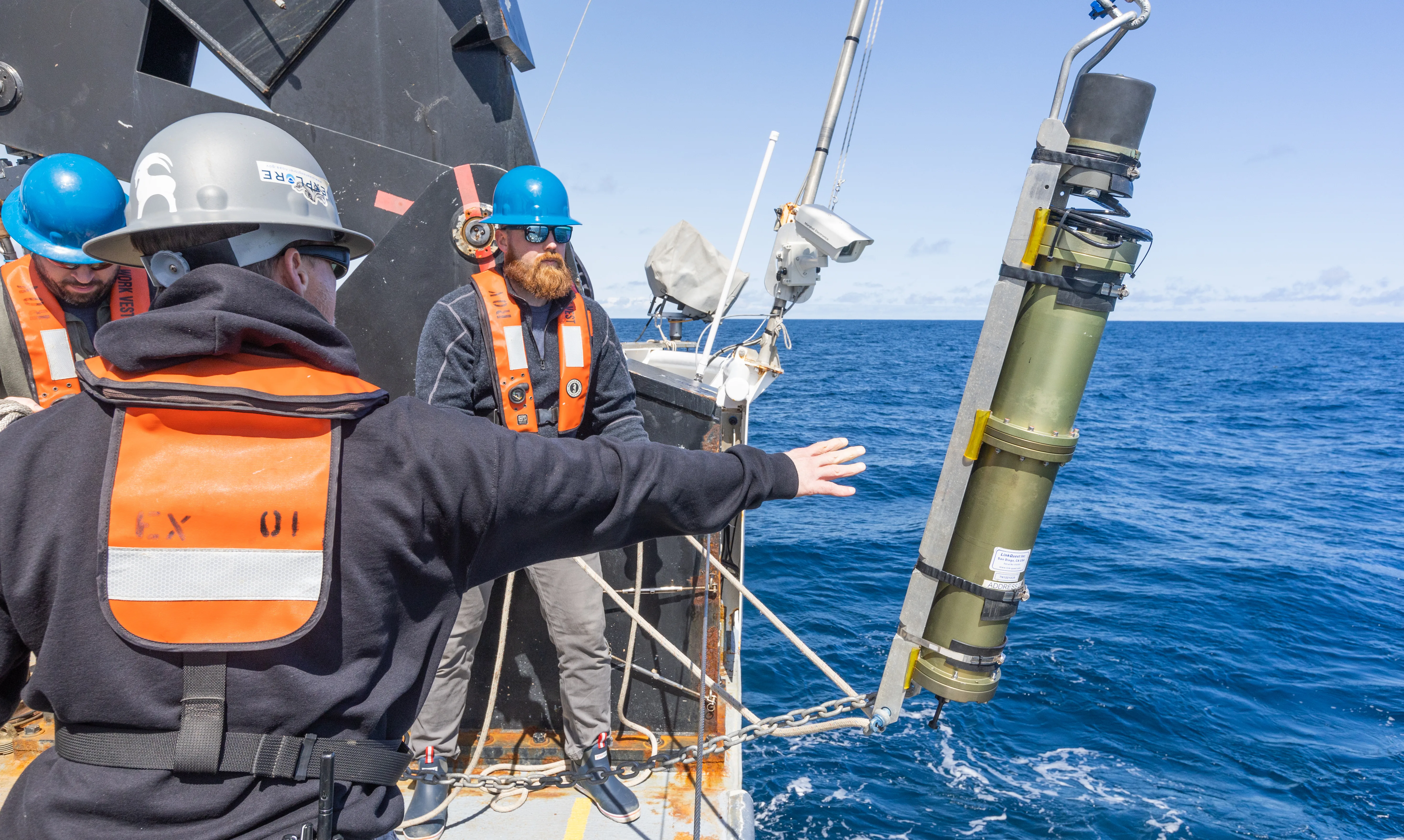 Crew on a ship deck preparing a long cylindrical USBL calibration transponder suspended beside the vessel