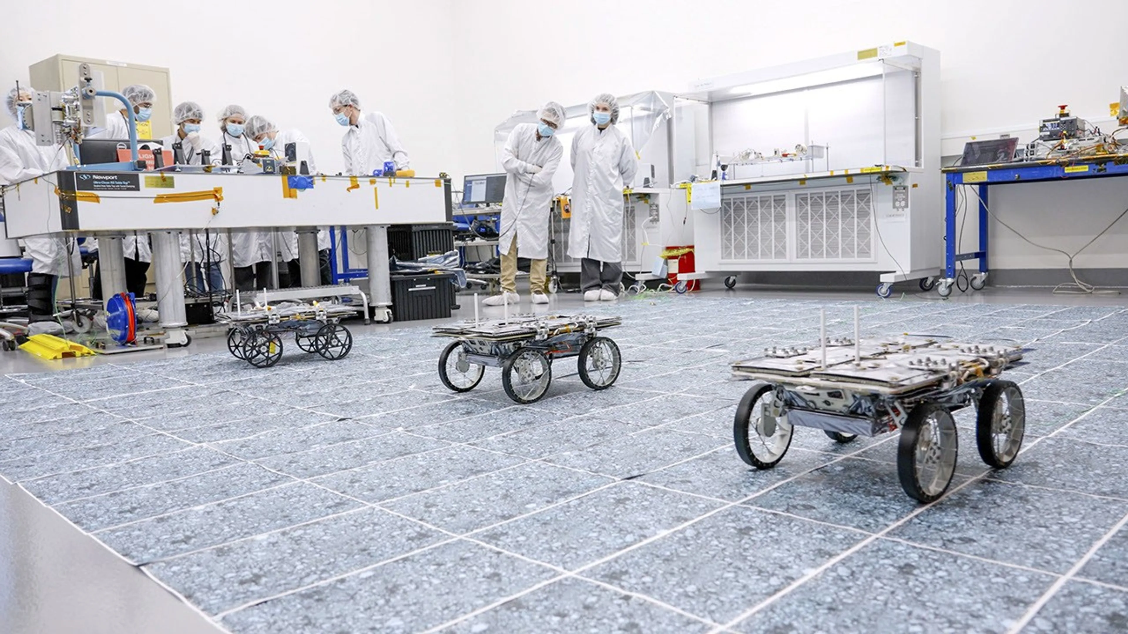 Three small four-wheeled CADRE rovers on a marked clean-room floor, with engineers observing from behind the test area.