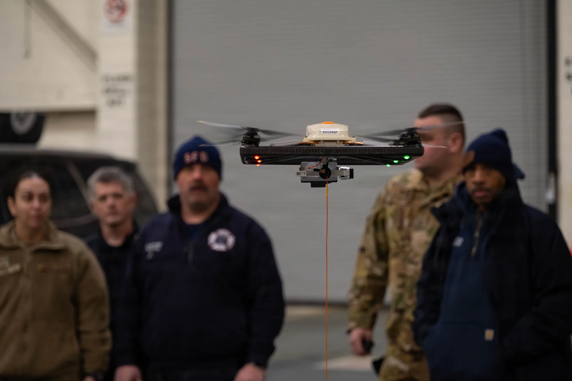 A quadrotor hovers indoors during training, with an orange tether descending beneath it and observers behind it.