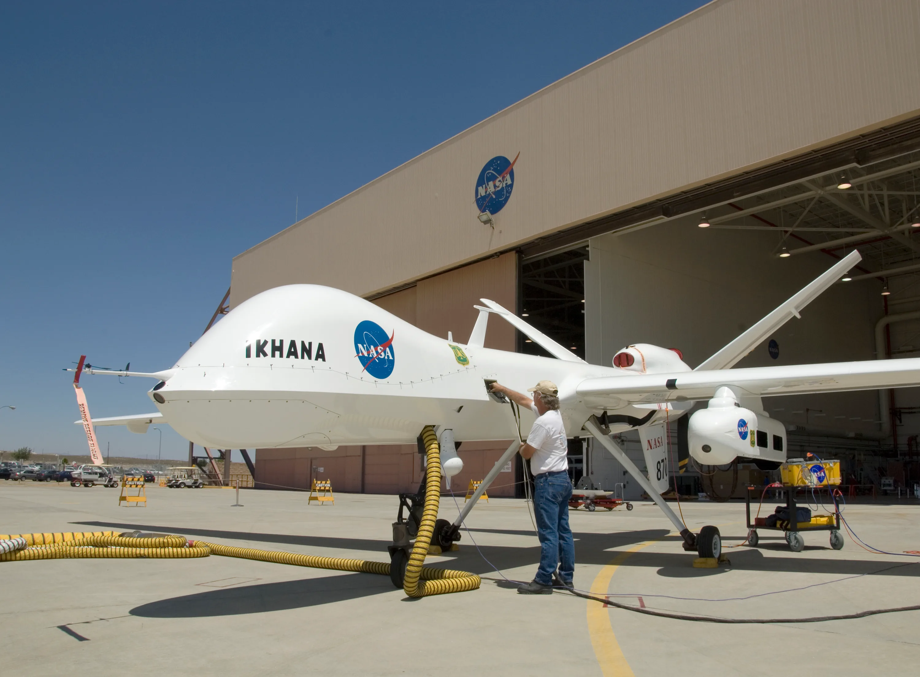 A ground crew member handles connections beside NASA Ikhana outside a hangar, with the aircraft nose, landing gear, and wing-mounted pod visible.