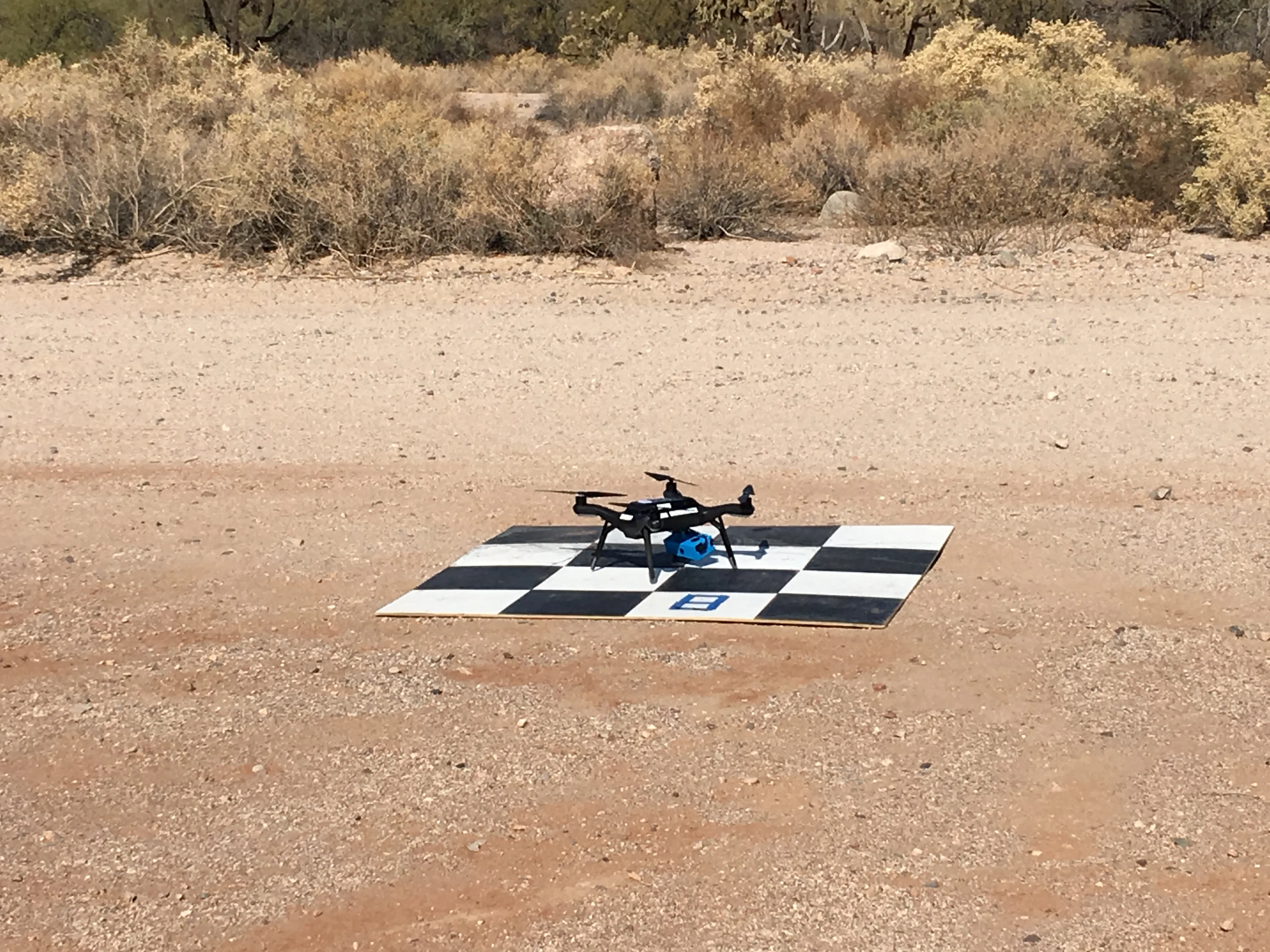 A small black quadcopter with a blue camera underneath rests on a black-and-white checked launch pad in dry terrain.