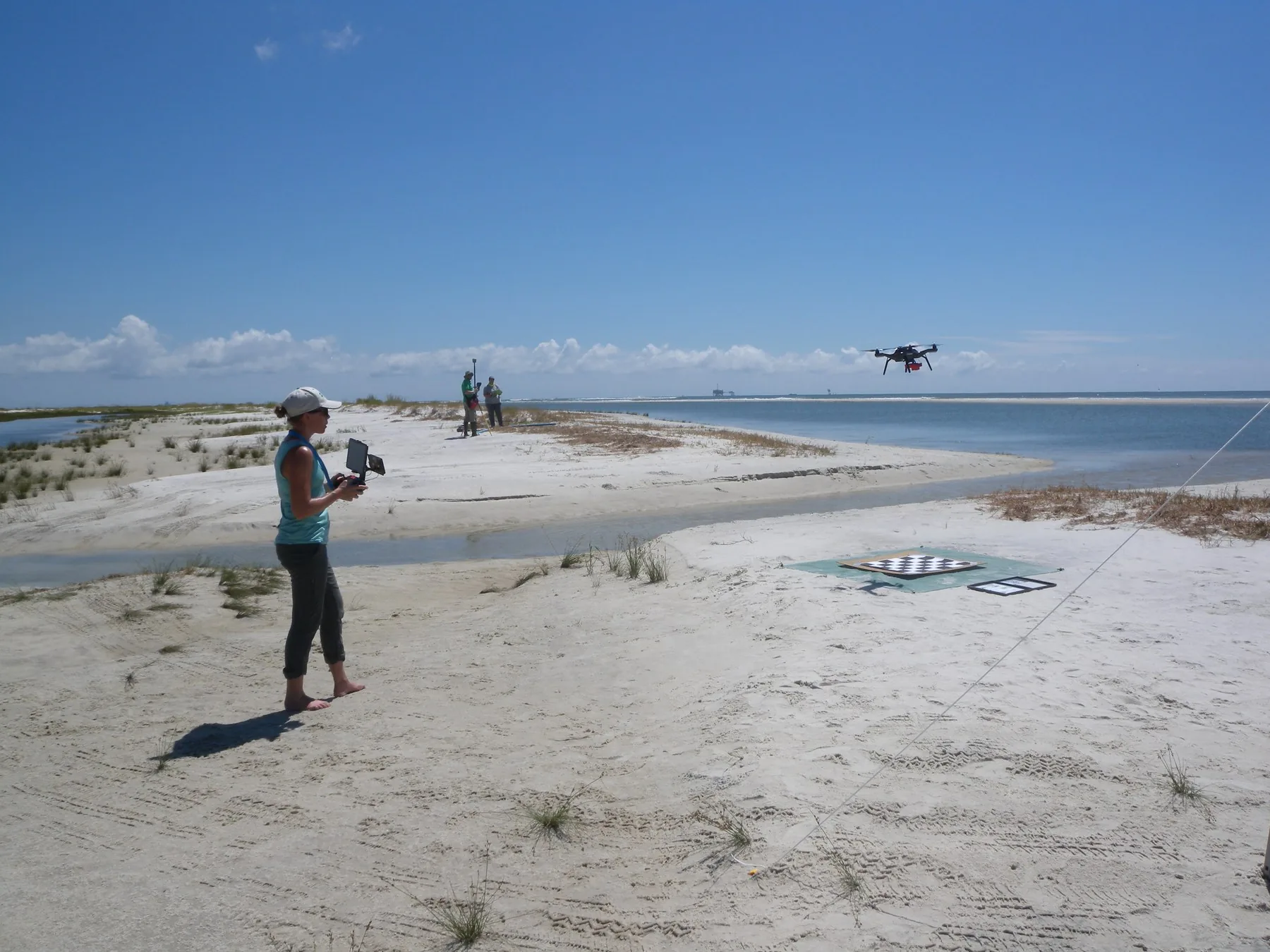 A pilot holds a controller while a multirotor aircraft flies above a beach with survey targets on the sand.