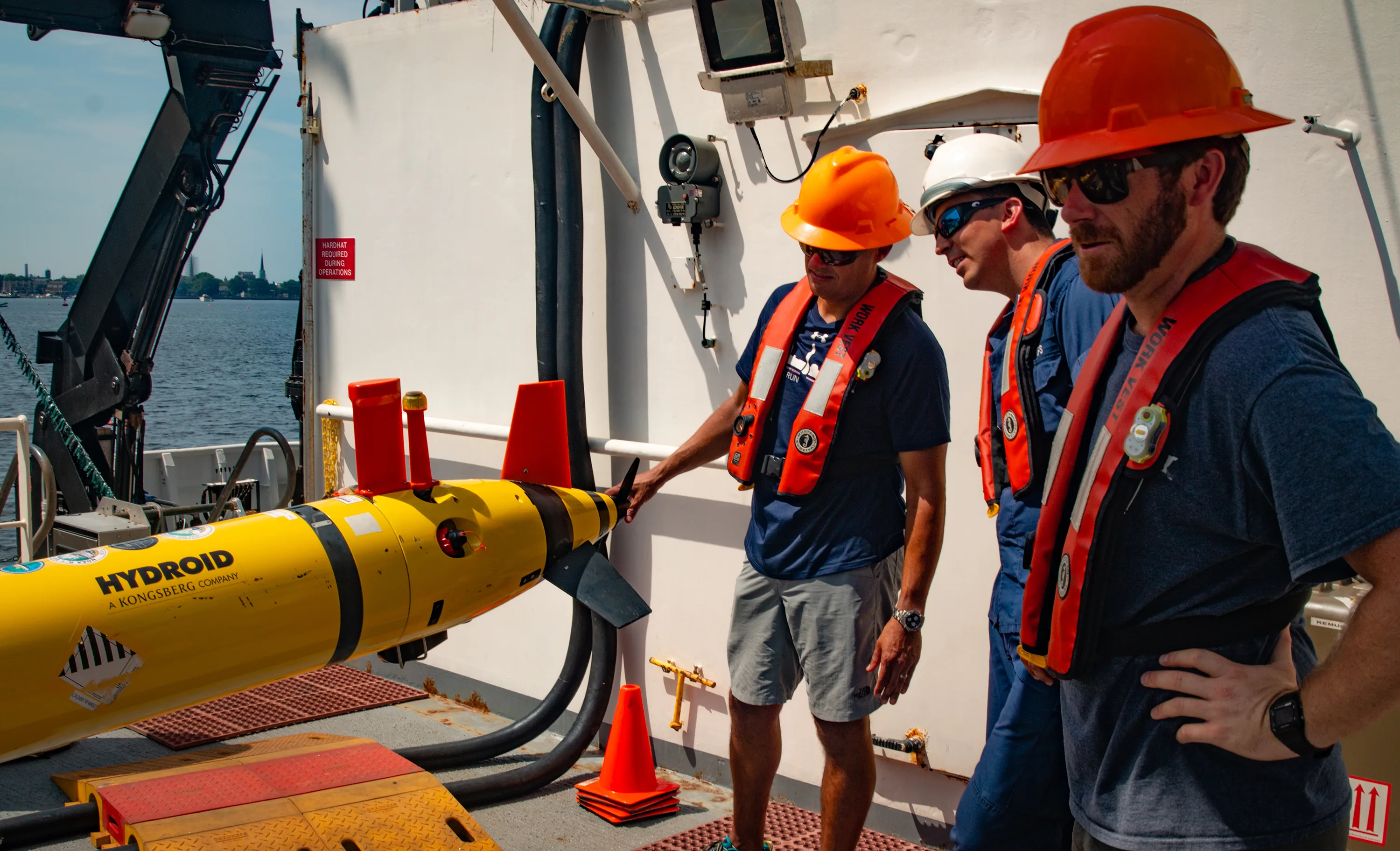 Three crew members in hard hats and flotation vests stand beside a yellow autonomous underwater vehicle on a ship deck.