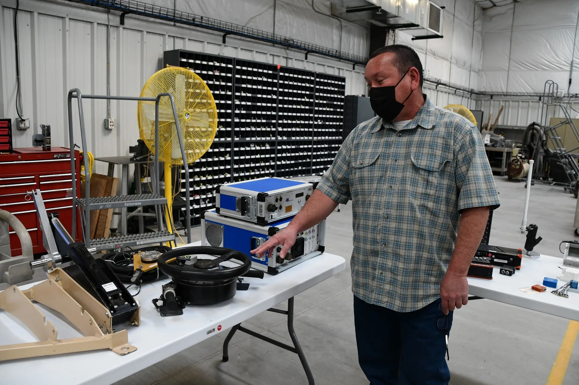 A technician points to a steering wheel and robotic vehicle-test equipment on a workshop bench