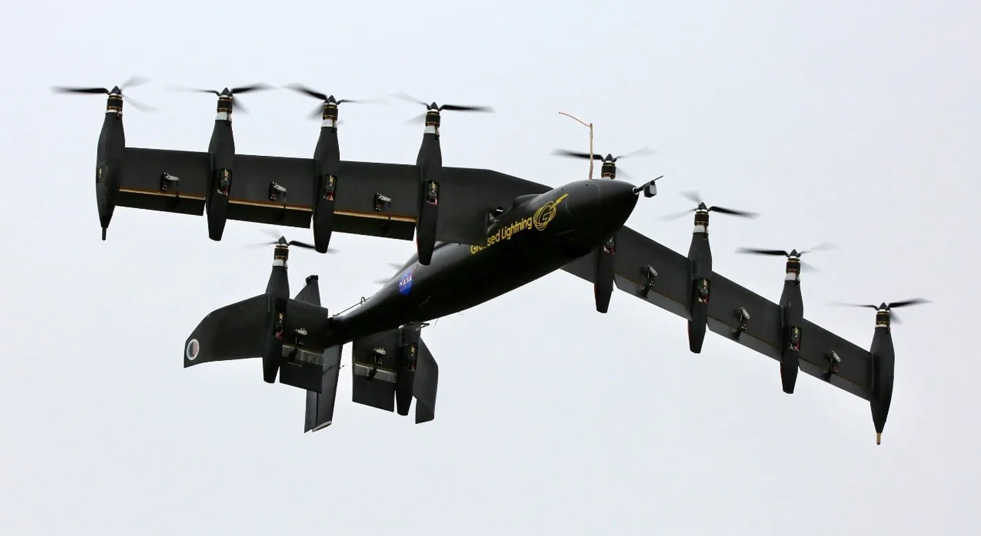 NASA Greased Lightning aircraft in the air, viewed from below with its wing and tail tilted and multiple propellers pointing upward.
