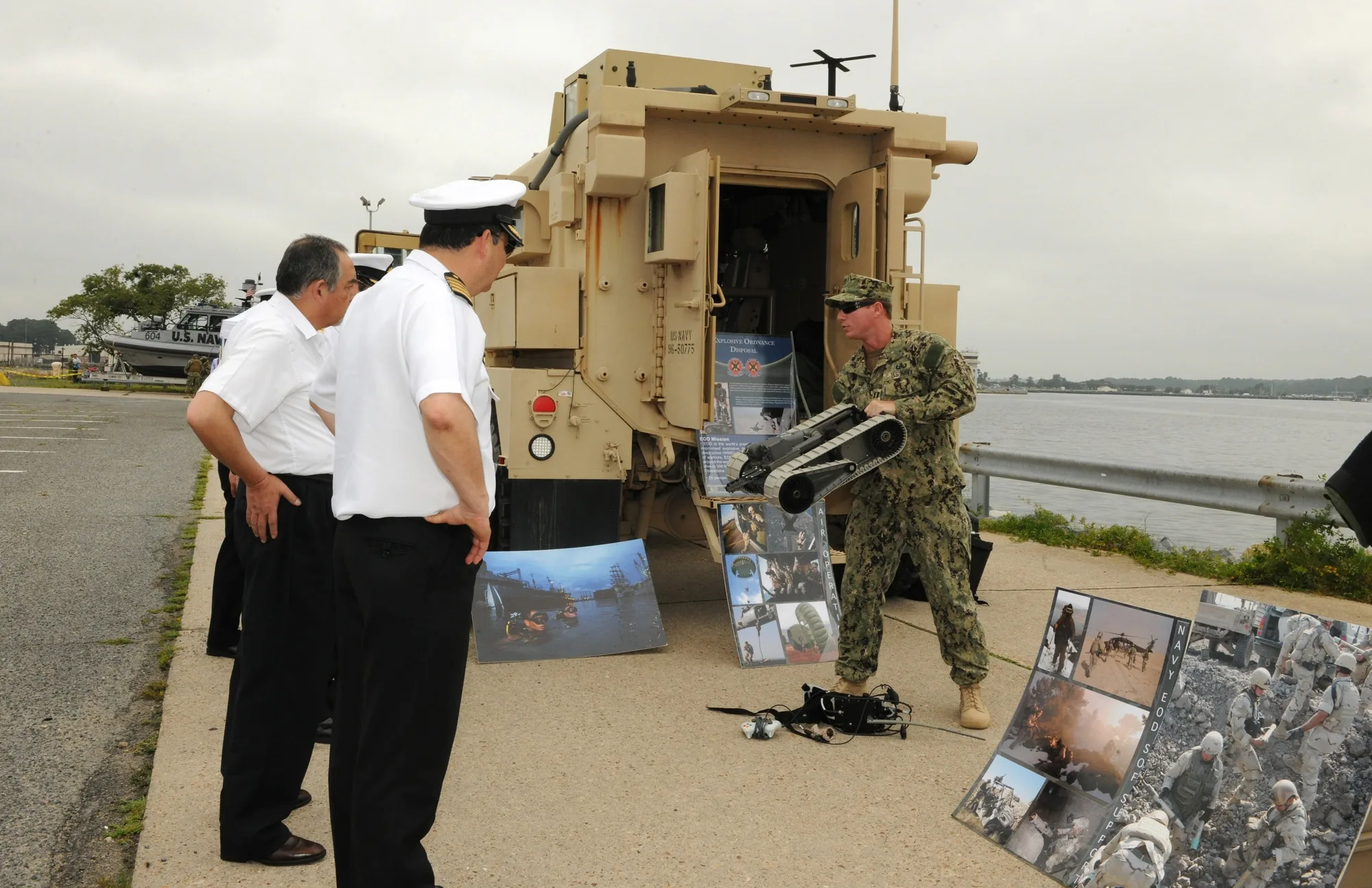 A sailor holds a compact tracked PackBot sideways during an outdoor demonstration, showing its continuous tracks to visitors.